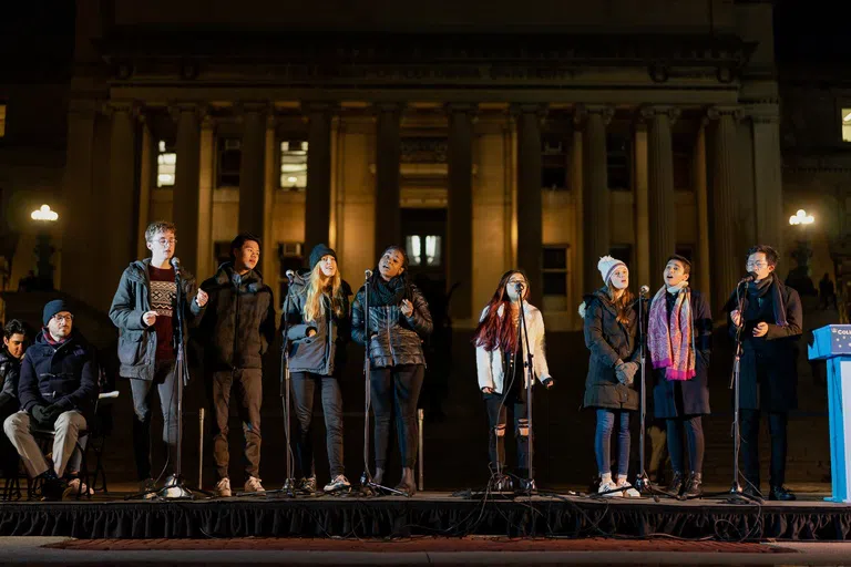 An a capella group of seven students stand on stage and sing, they sway, snap and are wearing winter clothes.