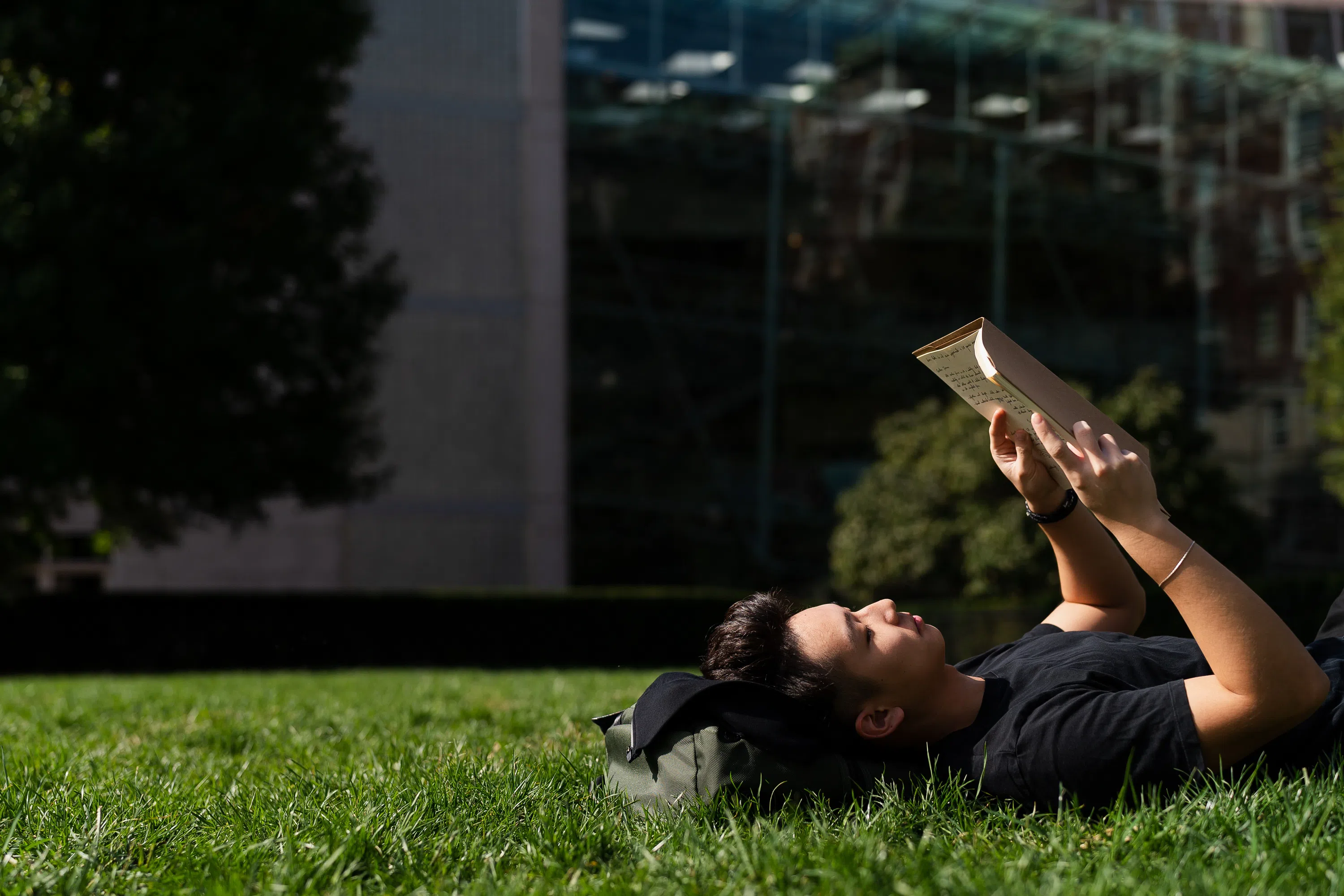 A student reads while laying face up in the grass and uses his bookbag as a pillow.