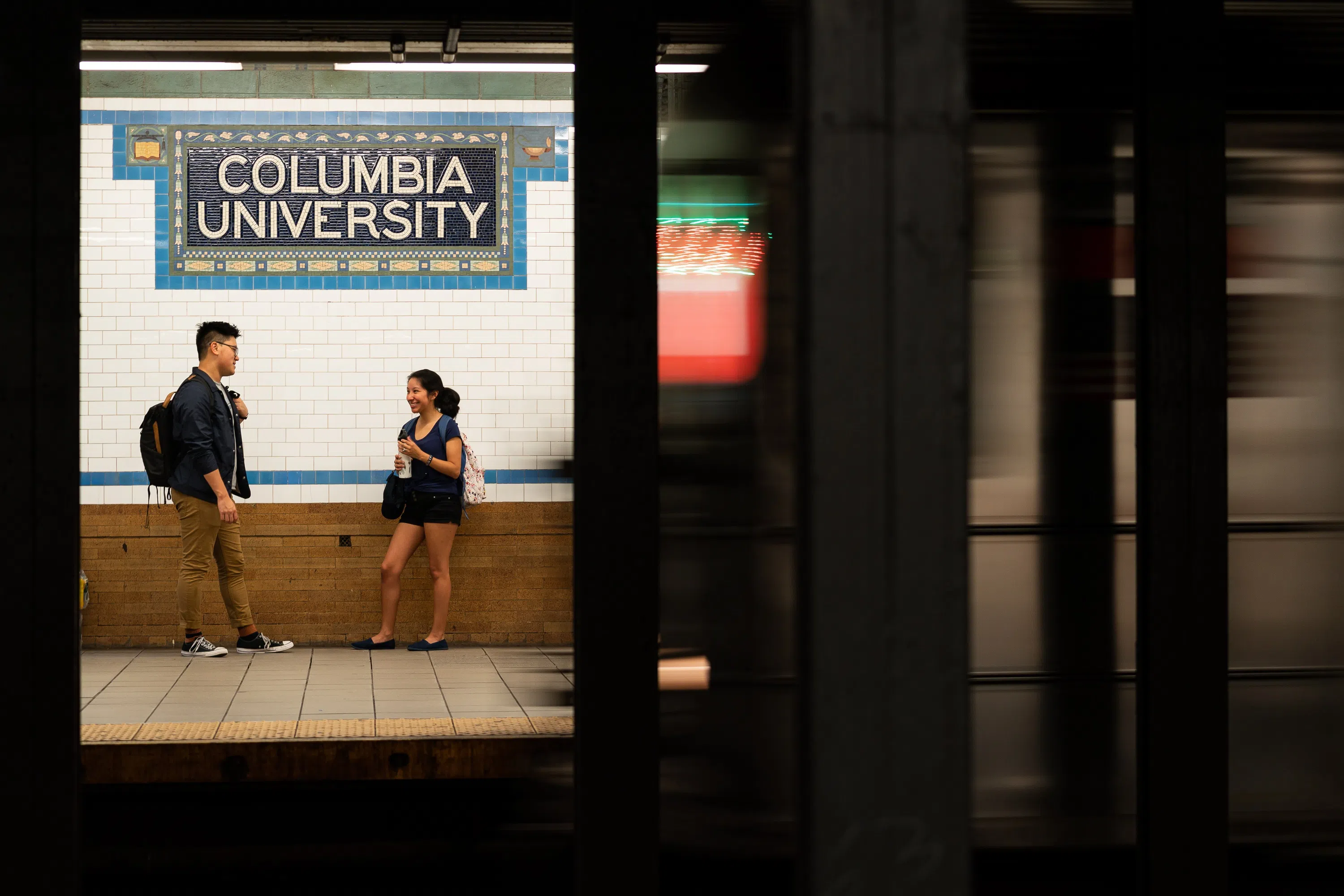 Two students stand on a subway platform as a train approaches; "Columbia University" is written on the wall in tile