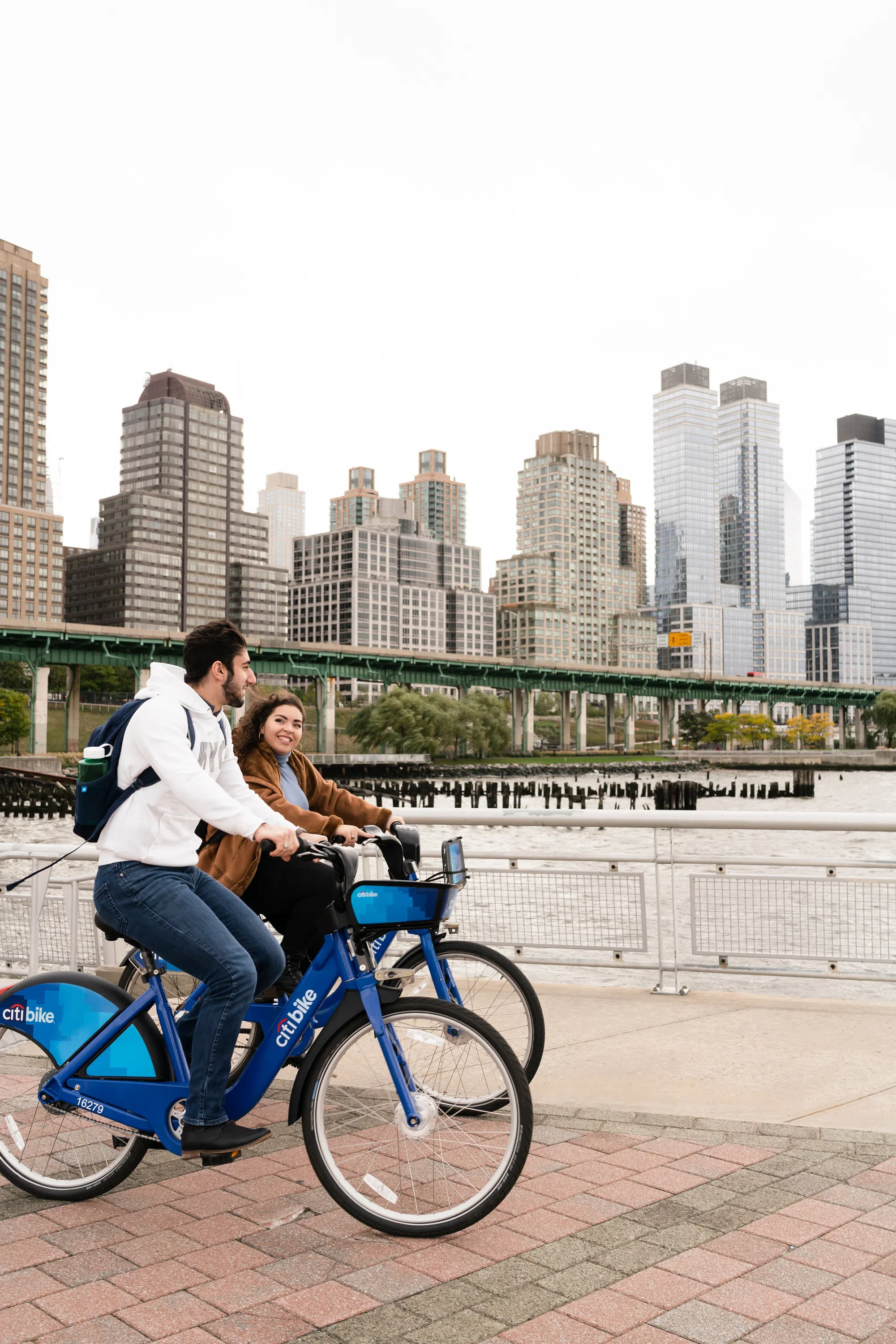 Two students ride bicycles down a park path. In the background is a body of water, a bridge, and a view of NYC skyscrapers.