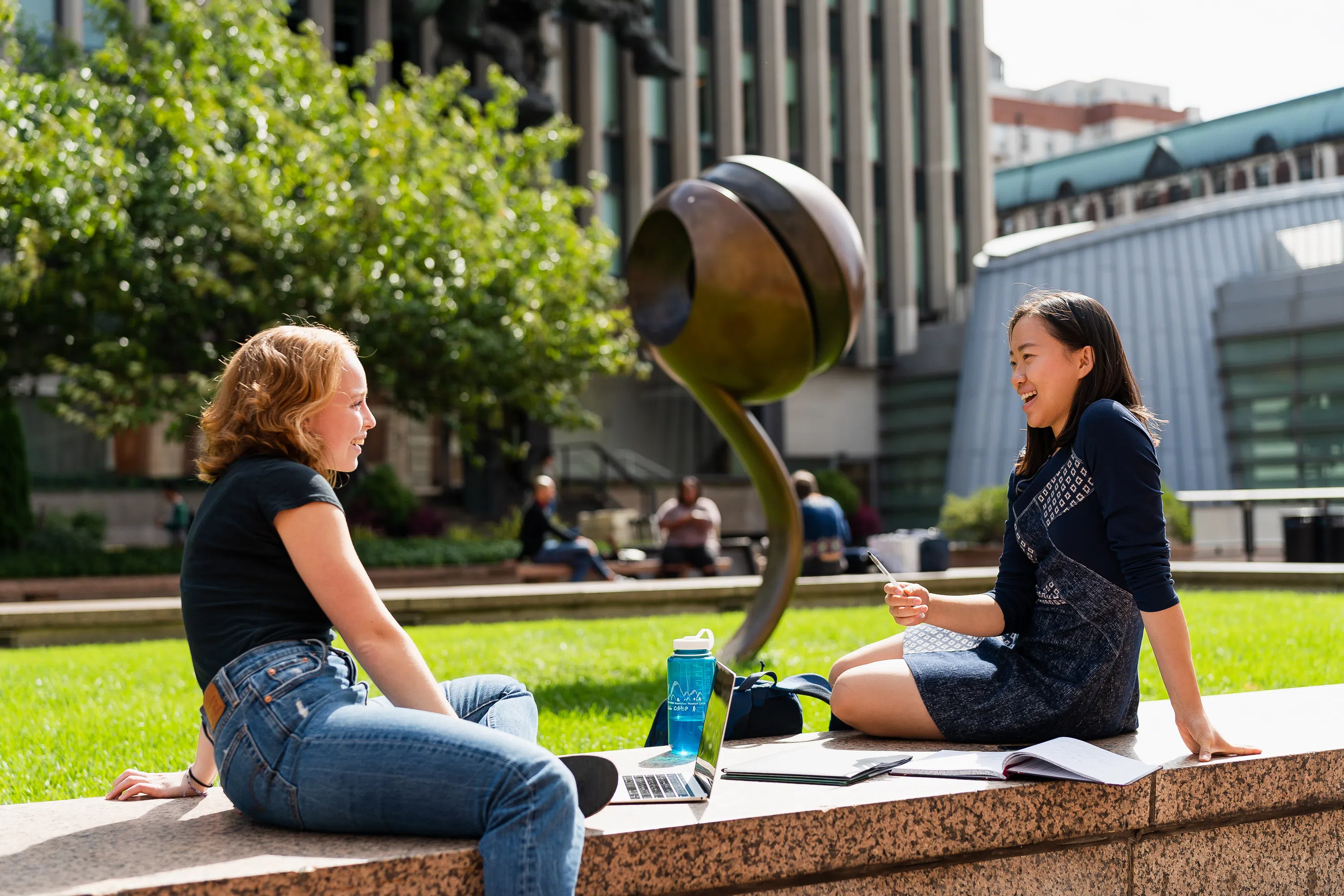 Two students lounge on a stone ledge while talking and laughing. They have books, laptops, and study materials in between them. Being them is sculpture art and lush trees.