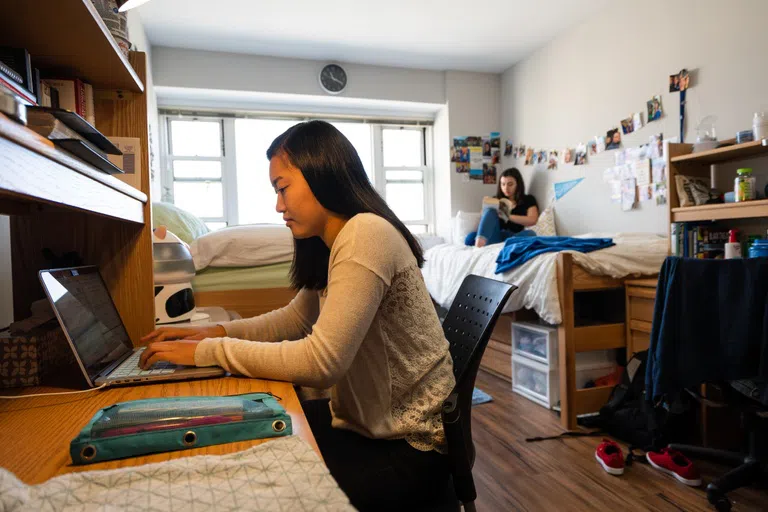 Two female students sit in a dorm room, one sits at her desk while the other is on the bed, both are studying.