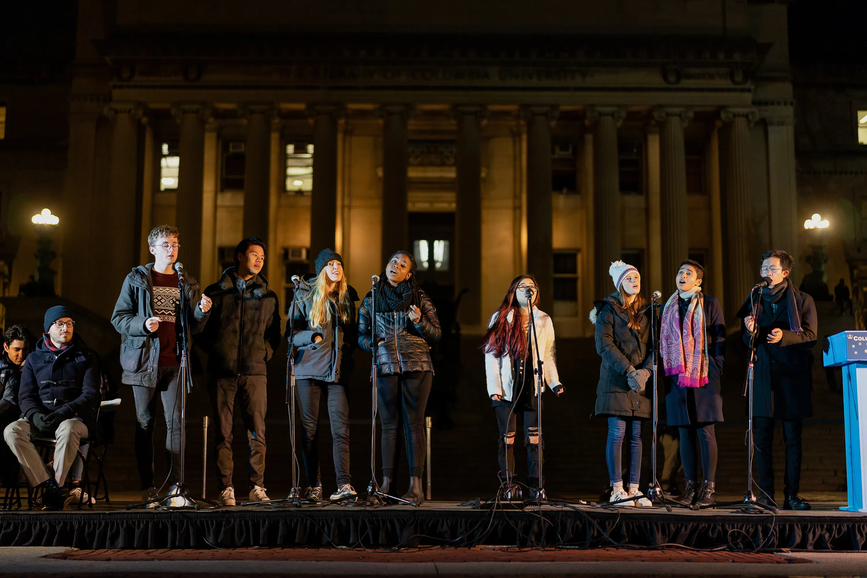 An a capella group stands on stage and sings, they are wearing winter clothes.