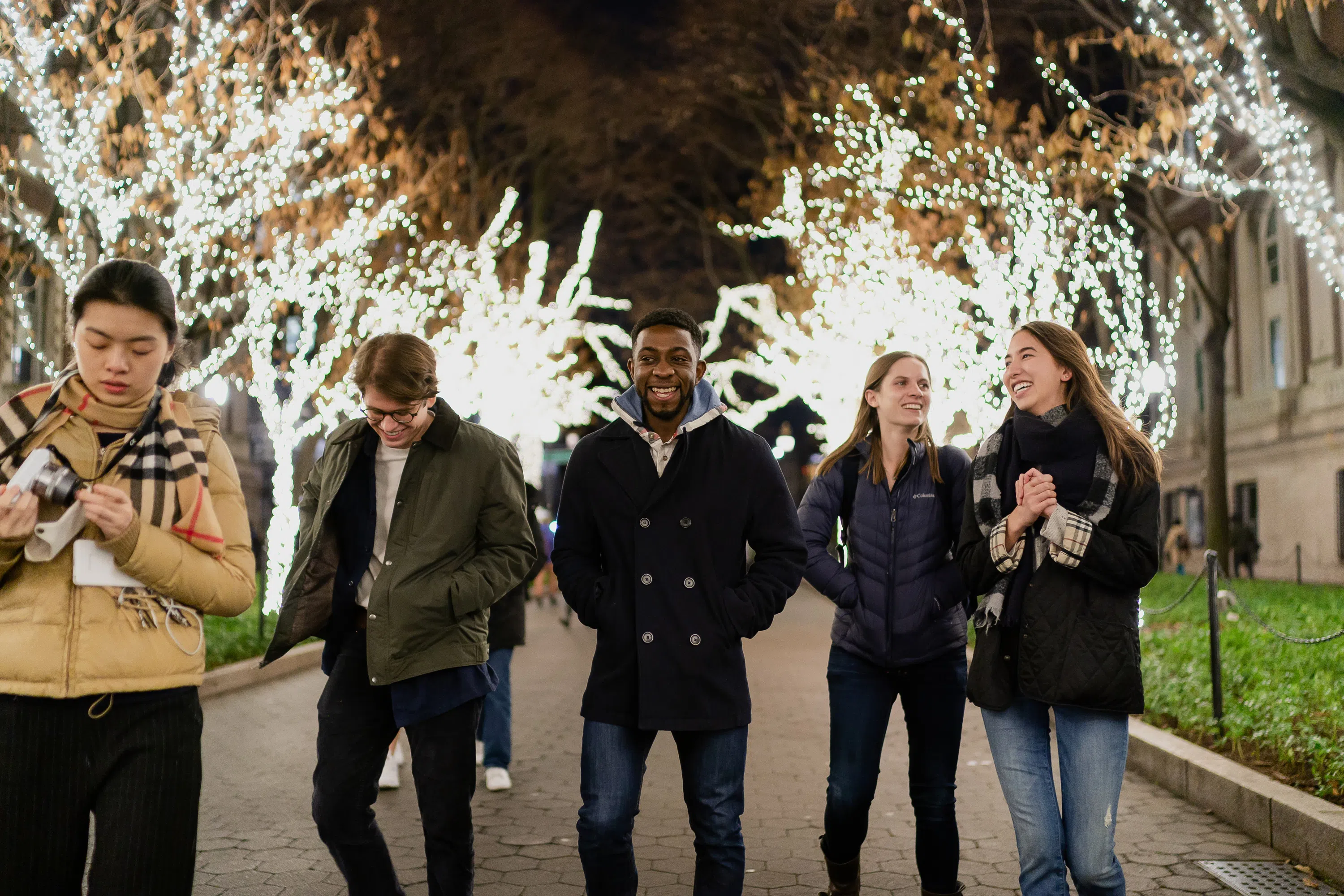 Four students walk forward while talking and laughing, the are surrounded by tress that are strung with brightly lit winter lights.