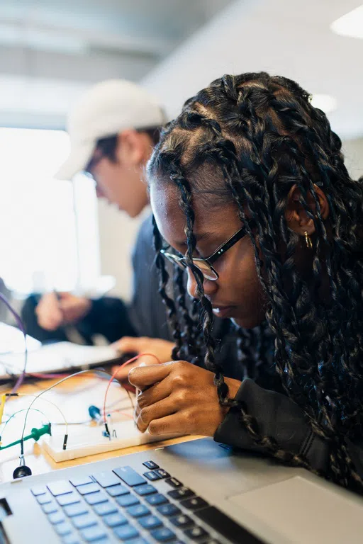 Female student tinkers with a small electrical wiring project with a laptop sitting in front of her.