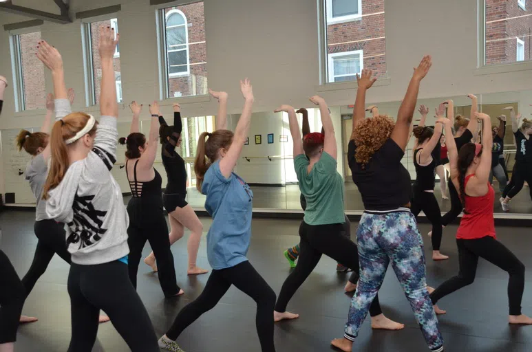 Students lined up in the dance studio, practicing positions. The studio has a wall of mirrors, dance bar, soothing white paint, and plenty of natural light