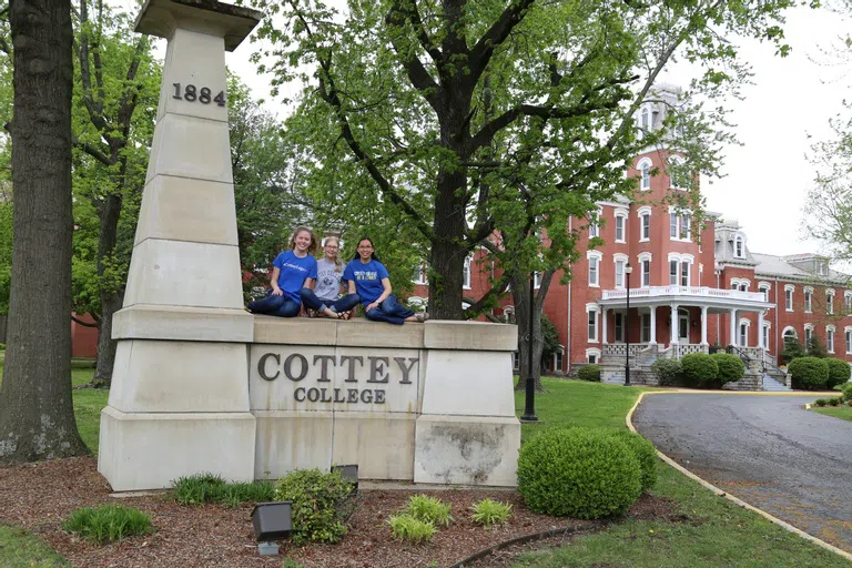 Three students sit happily on top of a stone sign that reads, "Cottey College." Main hall can be seen in the background