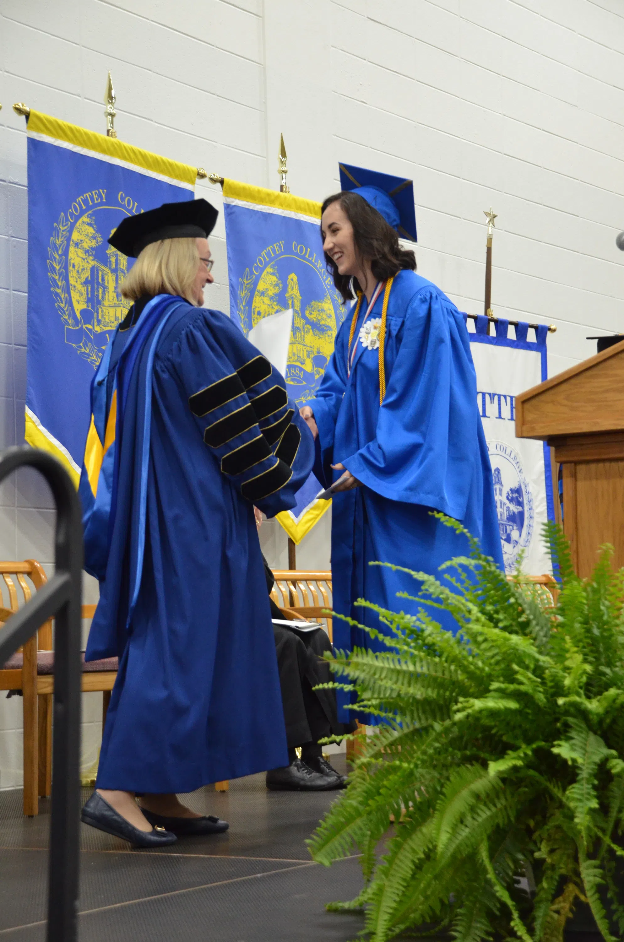 The president of the college, in full regalia, congratulates a new graduate with a smile and handshake