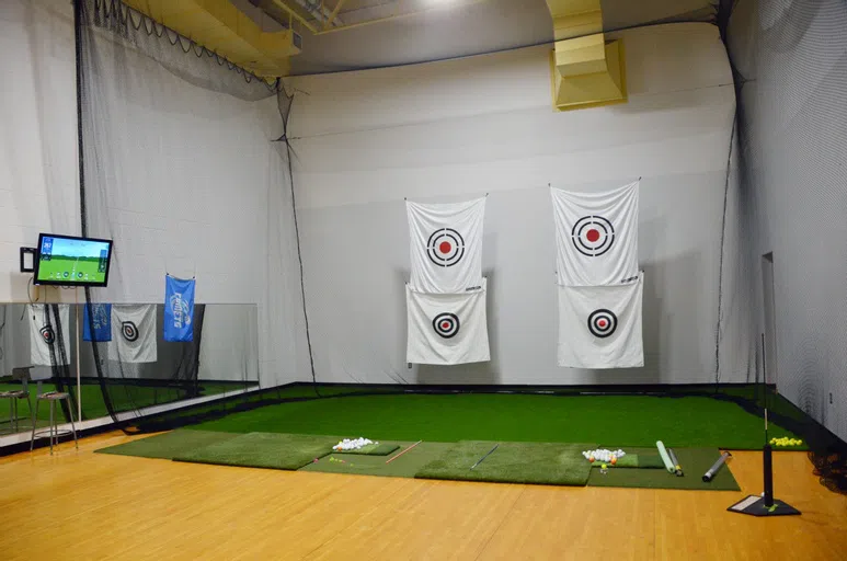 A large room with turf, targets, nets, golf balls, and a large screen to track your drive