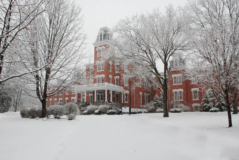 Snow is falling on Main Hall. Large snowflakes can be seen falling on the building and in the trees that populate the front lawn