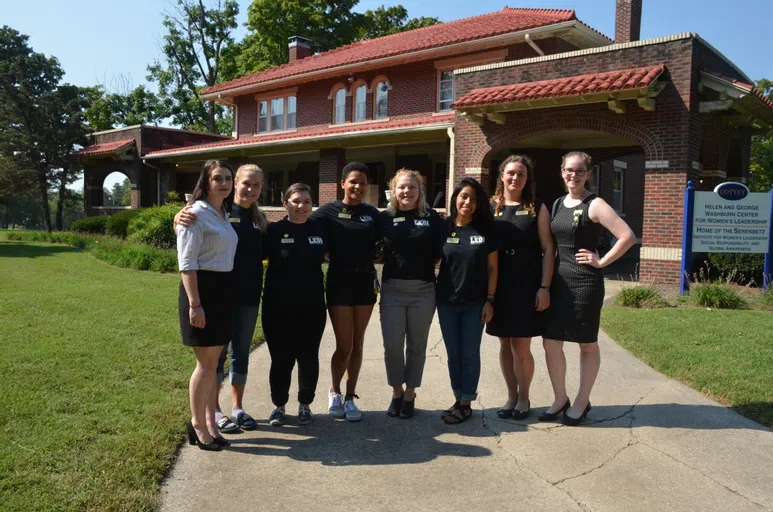 Eight female students standing proudly in front of a stately historical home