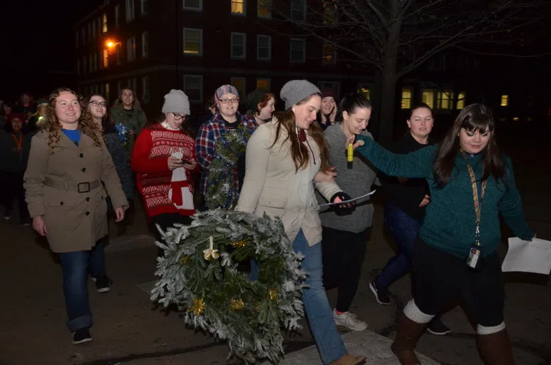 A group of students walking through the campus wearing sweaters and carrying wreaths. They are all looking down at the lyrics to the Christmas carol they plan to sing at their next stop