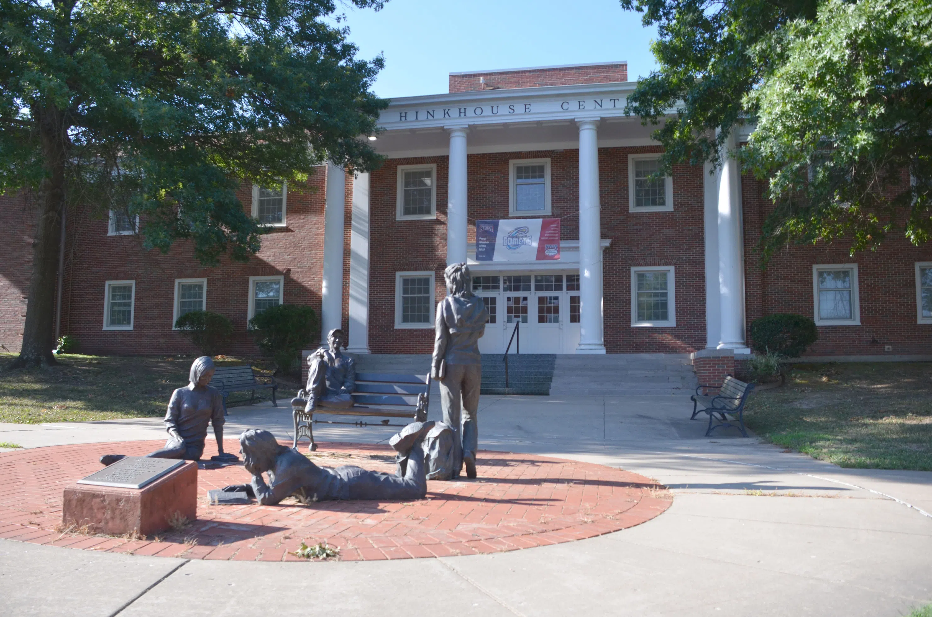 A stately brick building with large white columns; in the foreground is one of Cottey's most beloved pieces of art: four statues of students laughing, reading, and enjoying one another's company