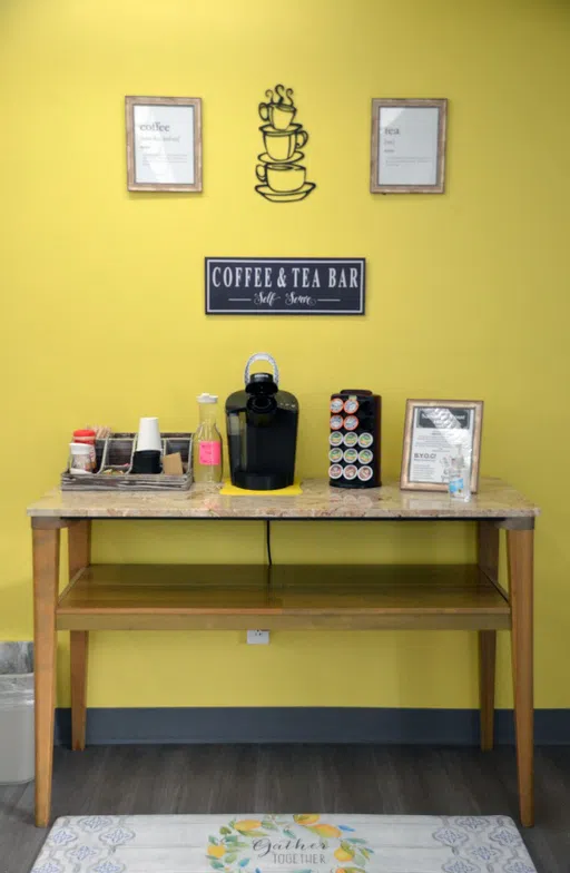 A wooden table holding a selection of coffees, teas, and the necessary equipment to make them, including cups, water, sweetener, and sugar. The wall behind the coffee station is Cottey yellow