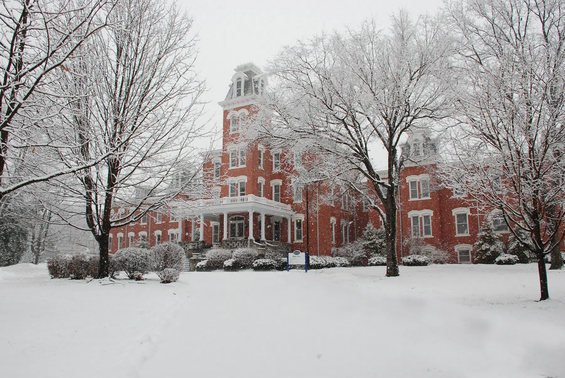 Snow is falling on Main Hall. Large snowflakes can be seen falling on the building and in the trees that populate the front lawn