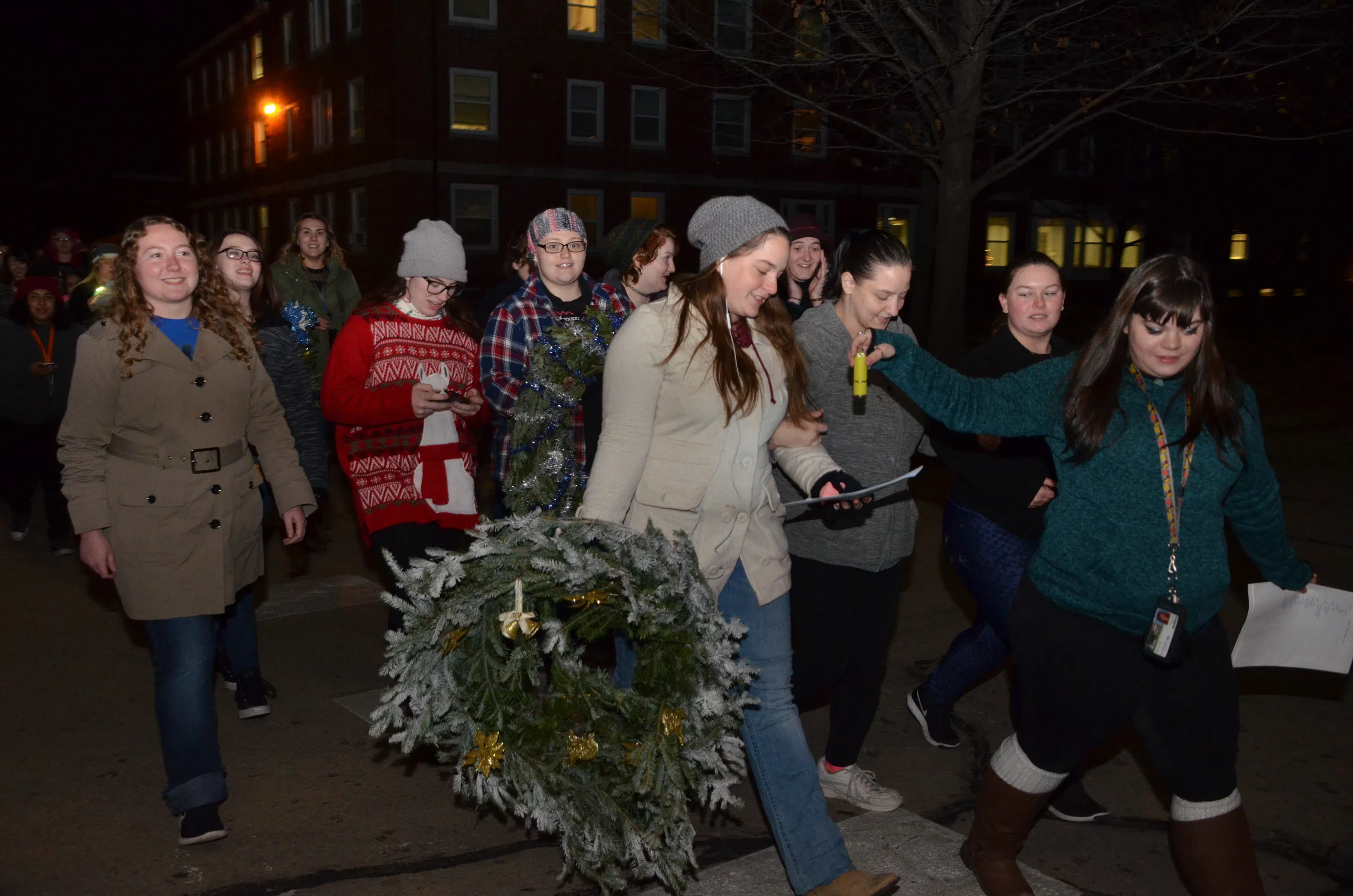 A group of students walking through the campus wearing sweaters and carrying wreaths. They are all looking down at the lyrics to the Christmas carol they plan to sing at their next stop