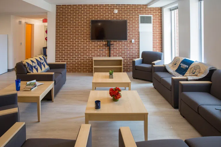 Lounge area inside residence hall with television, couches, tables and windows. 