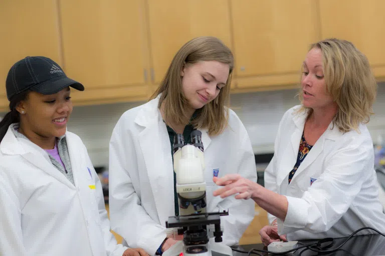 Two students in lab coats learn from a professor in a lab setting