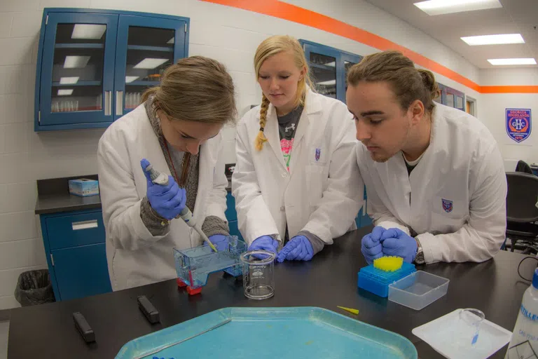 One student in a lab coat pipettes while two other students look on