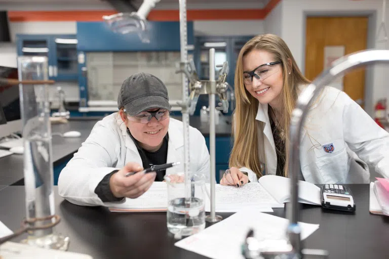 Two students in lab coats conduct an experiment. Both are smiling