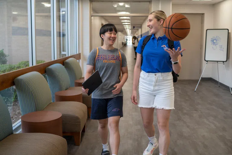 Two students walk down the hallway. Both are smiling. One is holding a basketball and one is holding a notebook. 