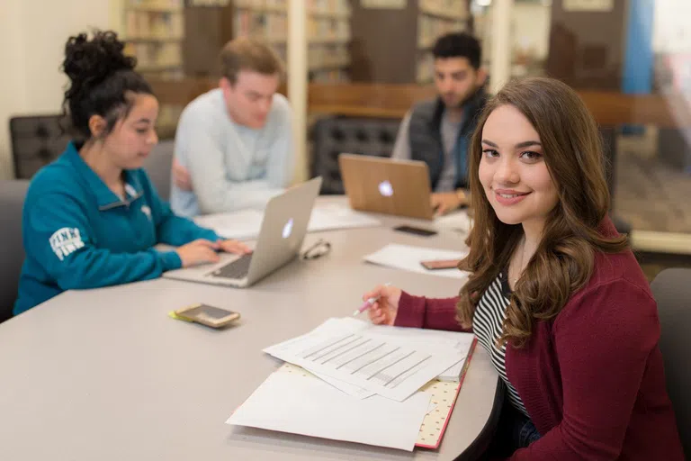 Students study at table. Nearest girl looks up from her papers to smile at the camera