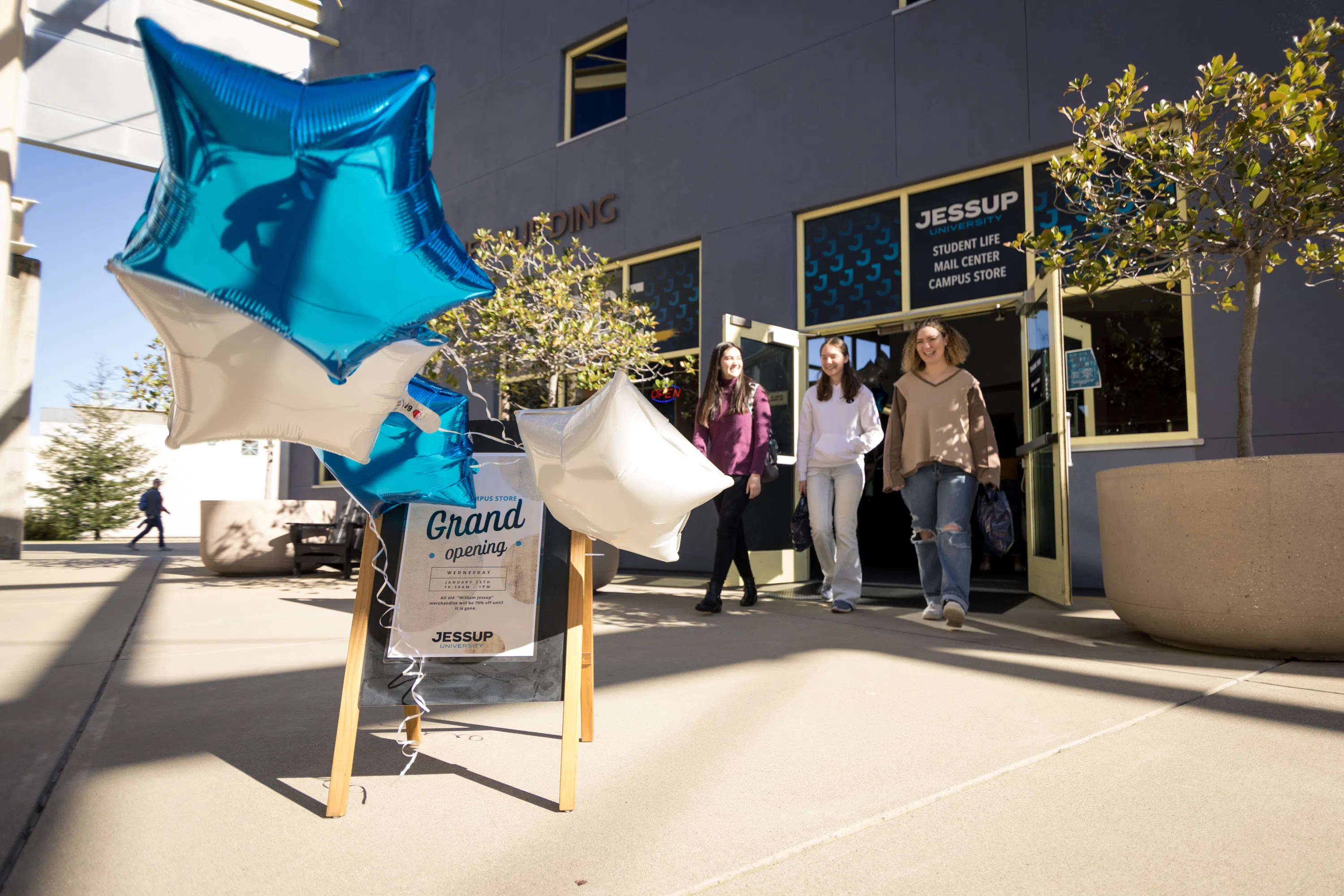 Three students exiting the Womack Student Life Building with a sign featuring balloons and "grand opening" text