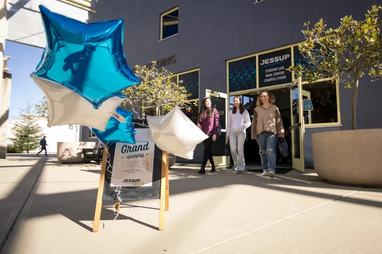 Three students exiting the Womack Student Life Building with a sign featuring balloons and "grand opening" text