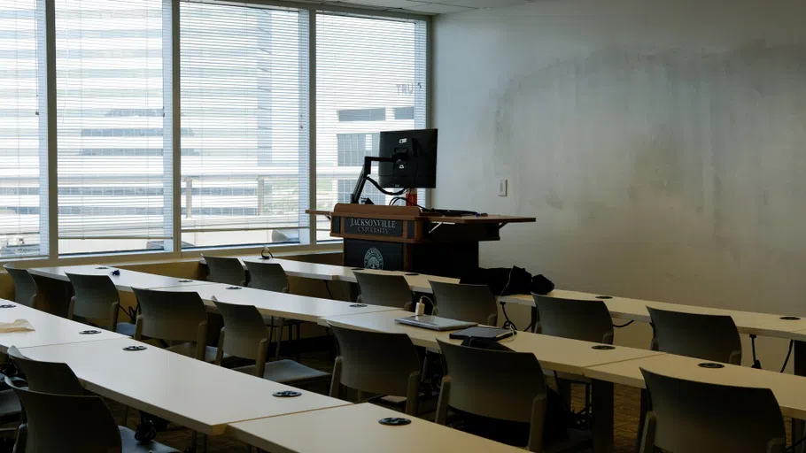 Multiple tables arranged in long lines, with a lectern and whiteboard at the front of the classroom. The left wall of the classroom is paneled with windows that provide a view of downtown Jacksonville.