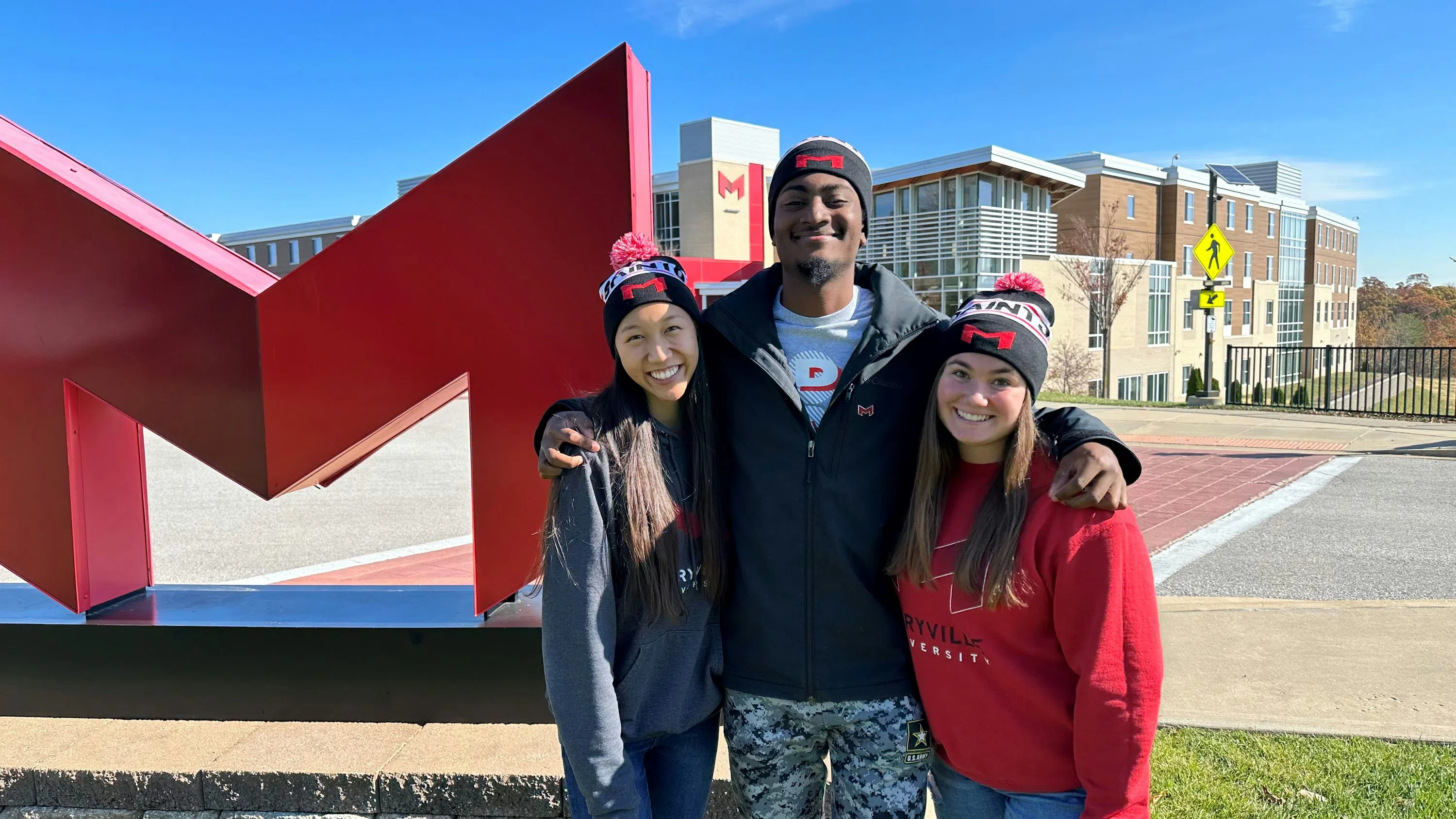 Three students stand in front of the Big Red "M" with Saints Hall in the background. 