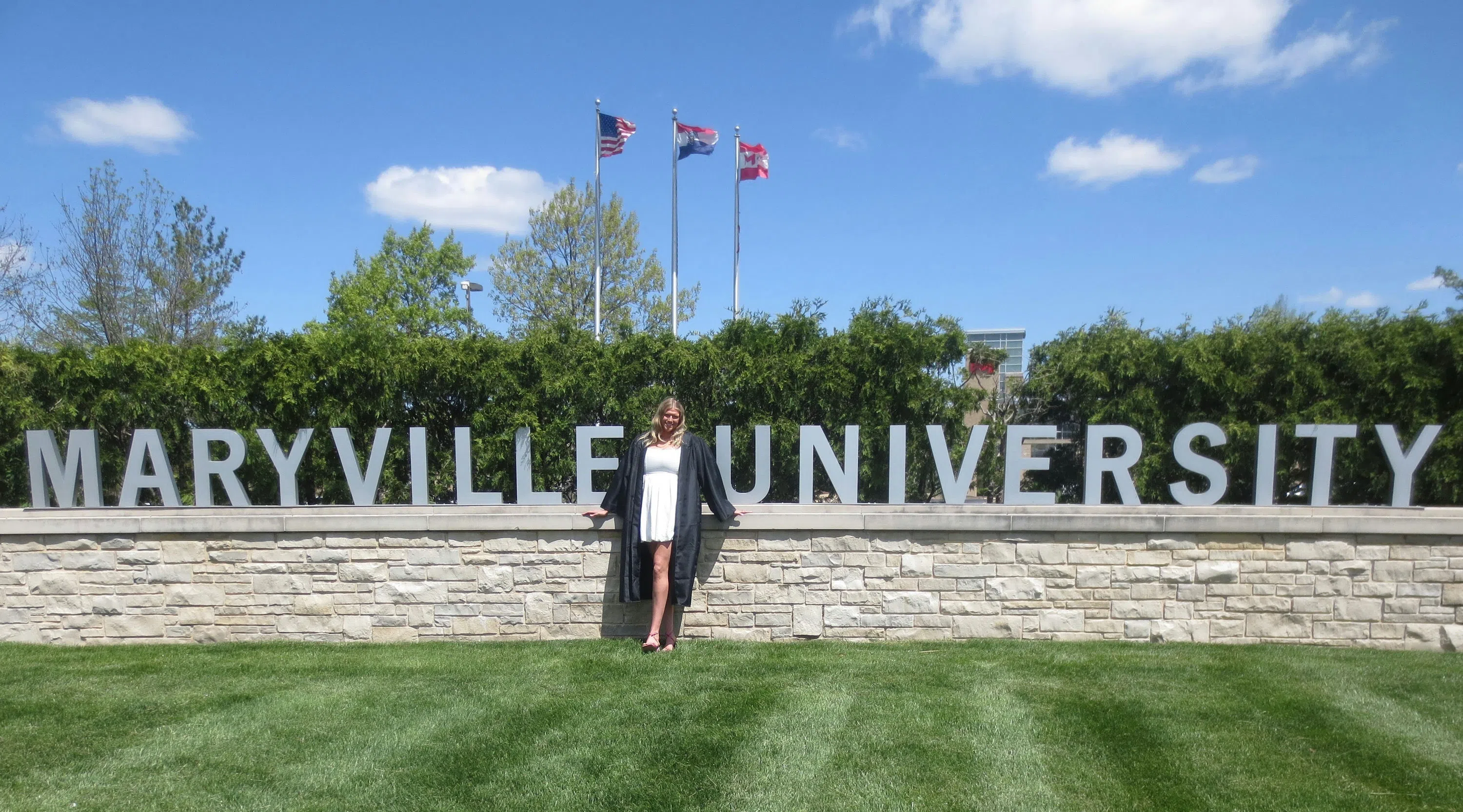 A student poses in their graduation cap and gown in front of the Maryville University sign.