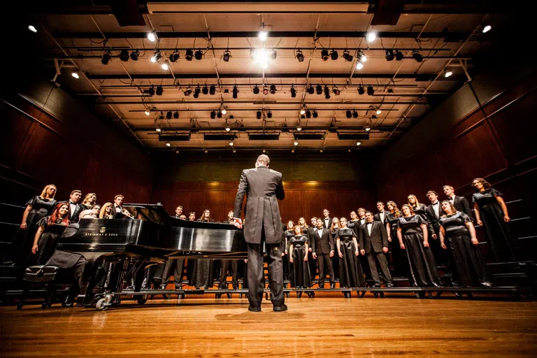 Conductor stands in front of a choir while they sing. The conductor's back is to the audience