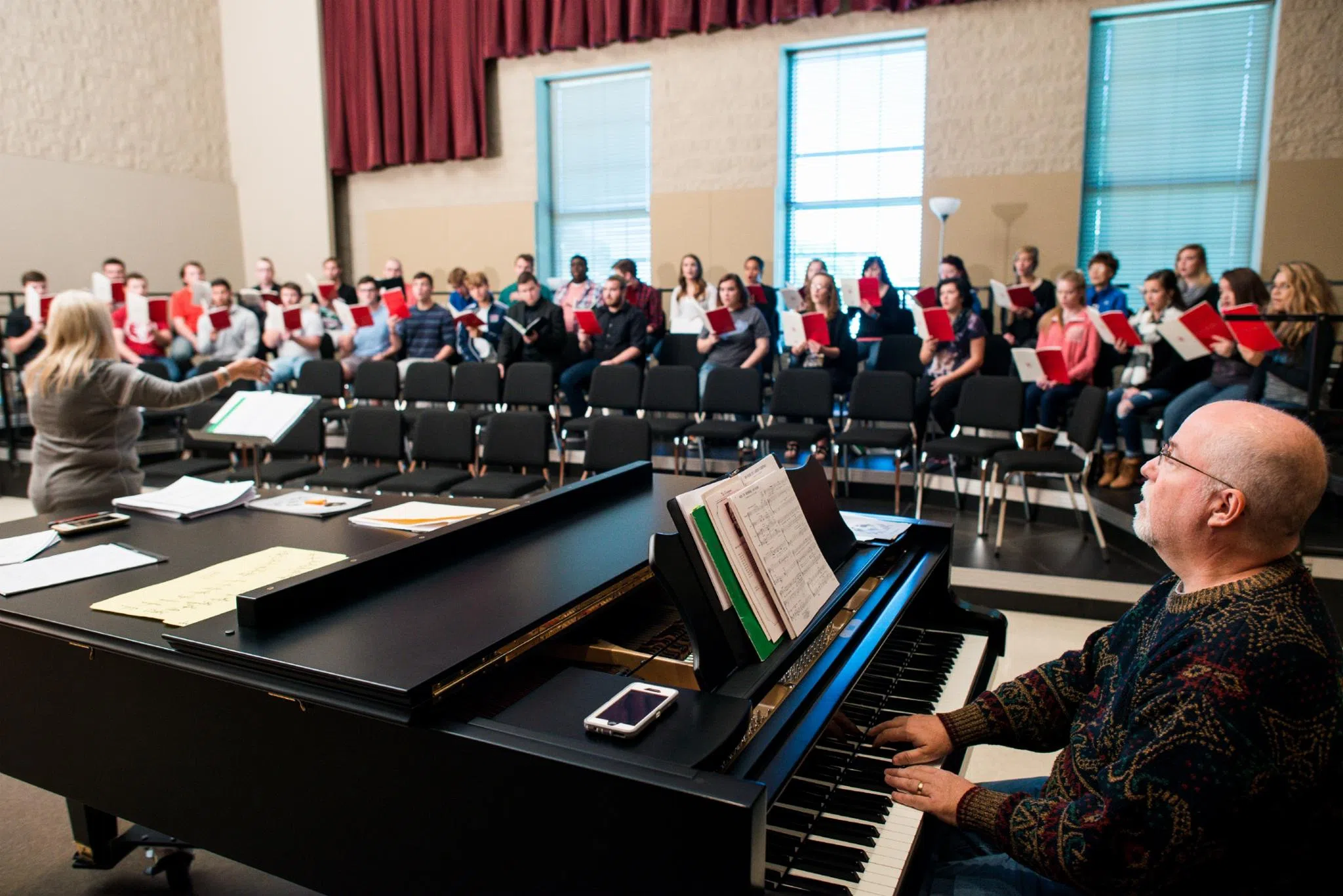 A pianist sits at a piano while the choir sits with their music books and rehearses 