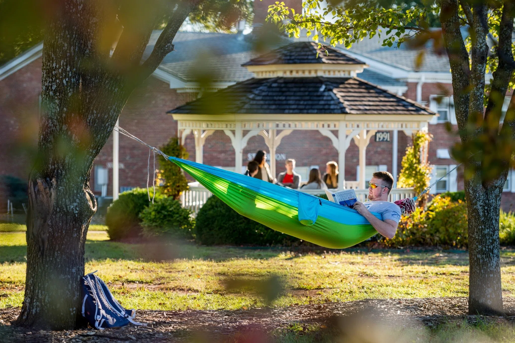Student hangs in a hammock on the Campus Mall. The gazebo stands nearby in the background, filled with students. 