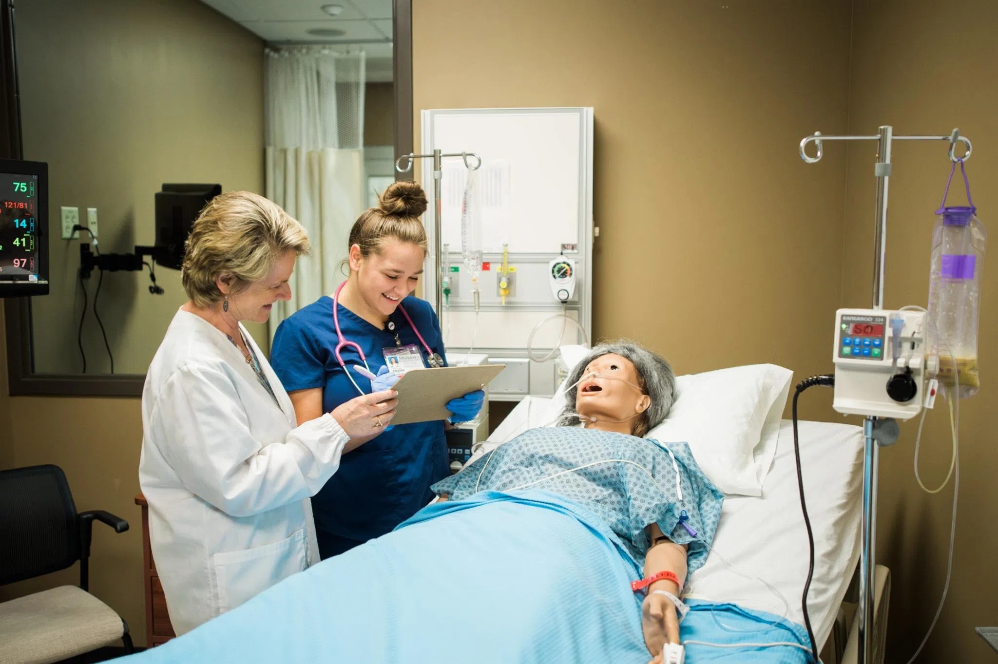Nursing student in scrubs looks at a clipboard with her professor while a manikin lays in the hospital bed. 