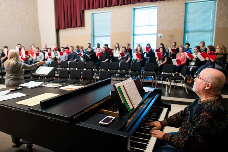 A pianist sits at a piano while the choir sits with their music books and rehearses 