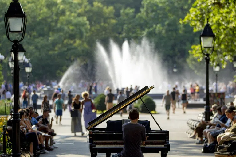 Man playing piano in Washington Square Park with water spouting from Washington Square Fountain in the background.