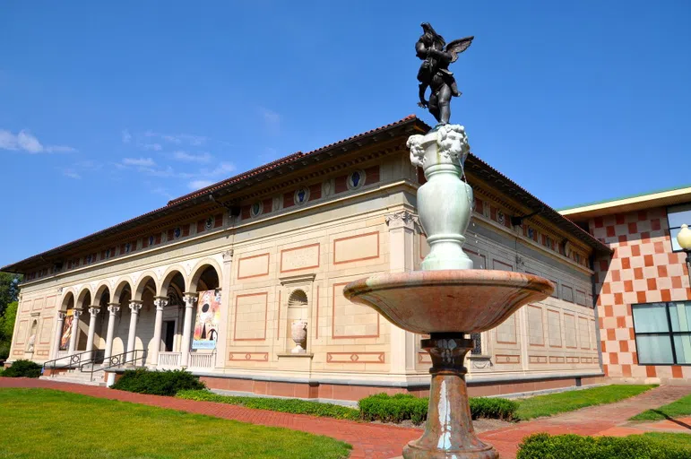 A cupid statue and fountain stand in front of a large, square tan building against a blue sky.