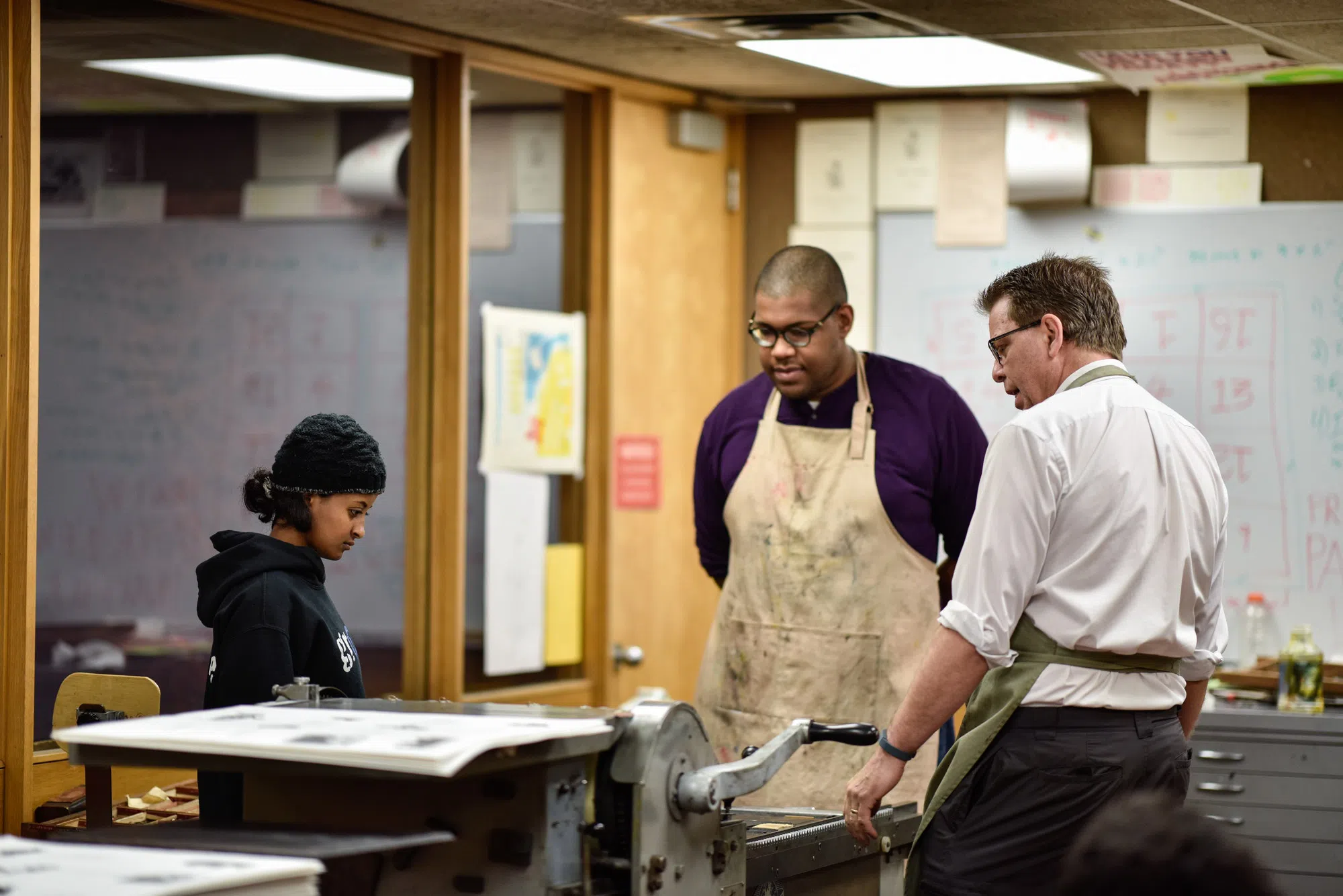 A professor and two students wearing aprons gather around the letter press. 