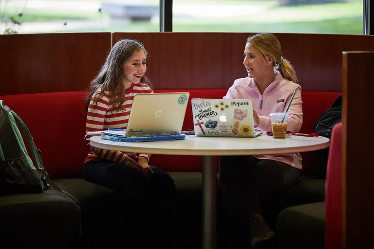 Two people on laptops working together sitting on red and black couches with a table in front of them