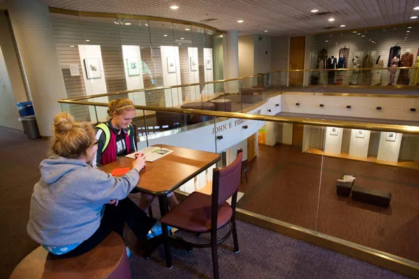 Two students sit at a table in an upper portion of a gallery. They are next to an area where you can look down into the lower floors.