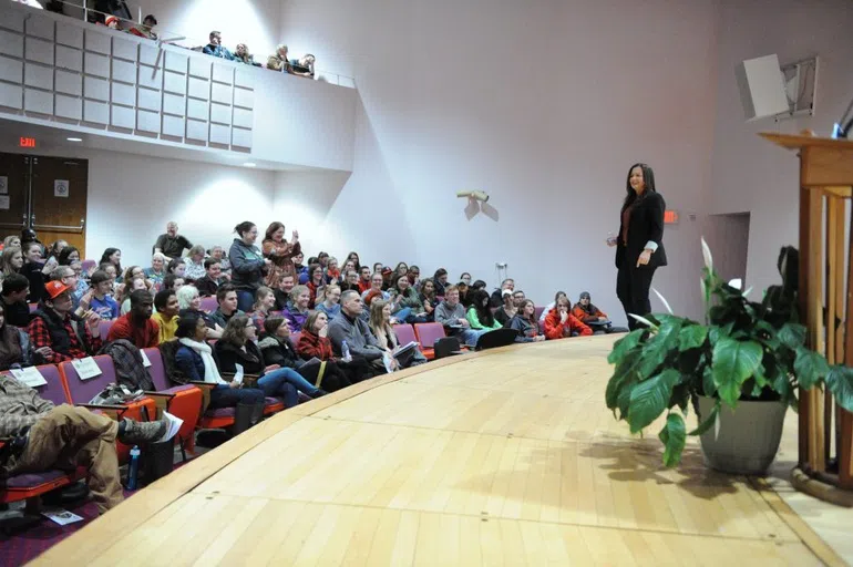 A woman stands on a stage amongst a group of people. They are listening to her speak.