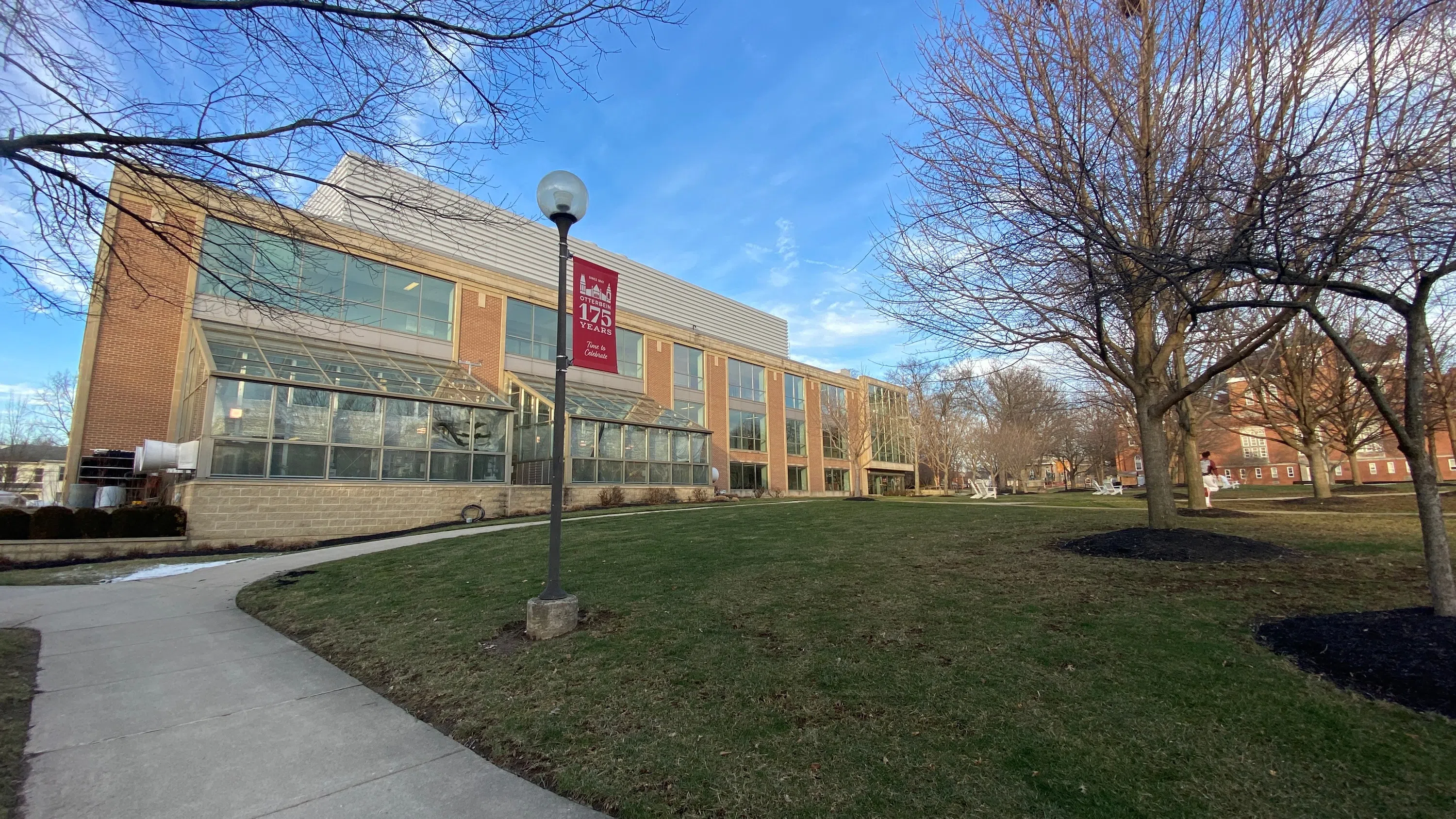 Light brown brick building with a glass greenhouse area in the front. The sky is blue with wispy clouds. There is a lamppost in the front with a 175th banner.