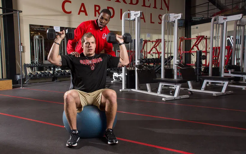 Man lifting weights with a male trainer behind him.