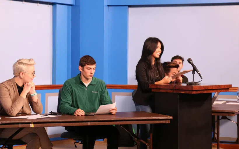 A woman is speaking at a podium in a classroom setting amongst a few other colleagues