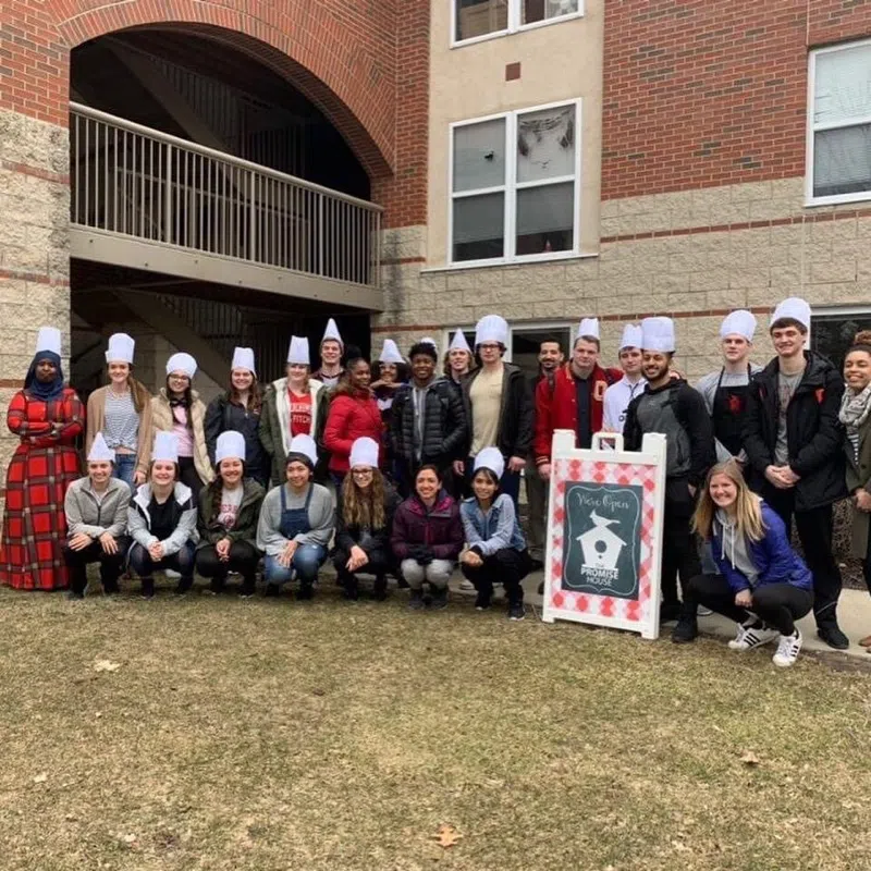 A large group of students wearing chef hats pose together outside the Promise House.