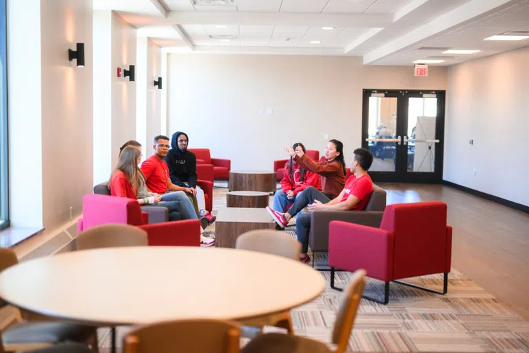 A lounge area with single red plush chairs and light beige tables. There are people conversing at one set of chairs