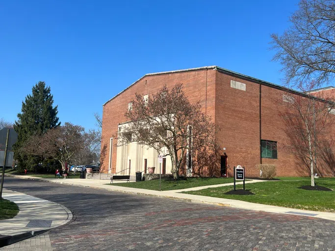 A brick building with beige entryway sits behind trees. 