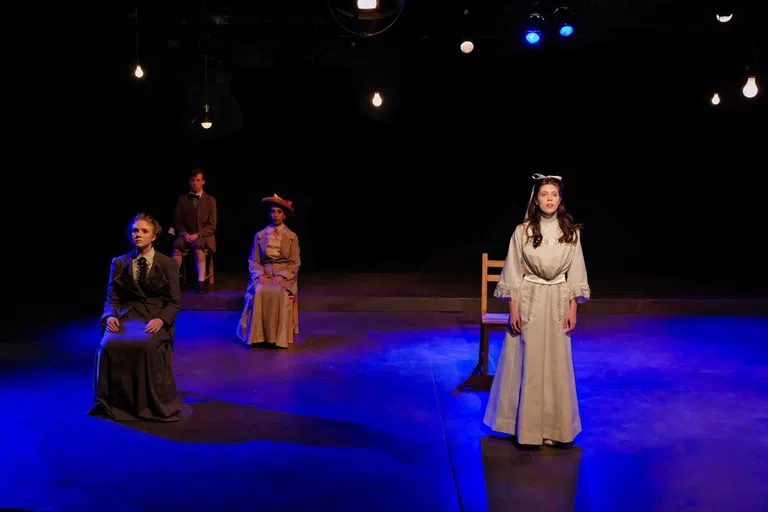 4 people are on a blue-lit stage. A woman is standing and performing to the crowd, and the other three are sitting in chairs looking out into the audience