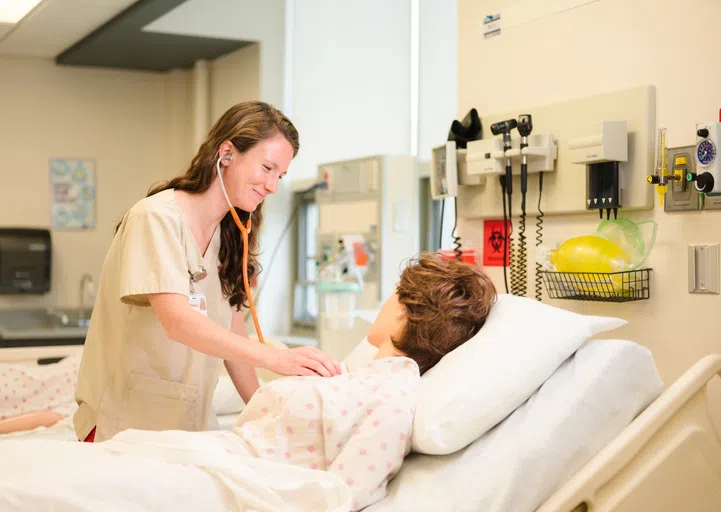 A woman is performing care on a practice dummy in a hospital lab setting.